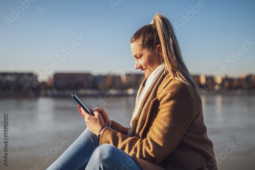 Beautiful woman in warm clothing using mobile phone and enjoys resting by the river on a sunny winter day. Toned image.