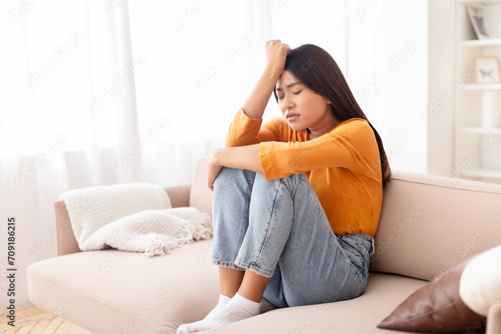 Upset young asian woman sitting on couch, touching her head Upset young asian woman sitting on couch, touching her head