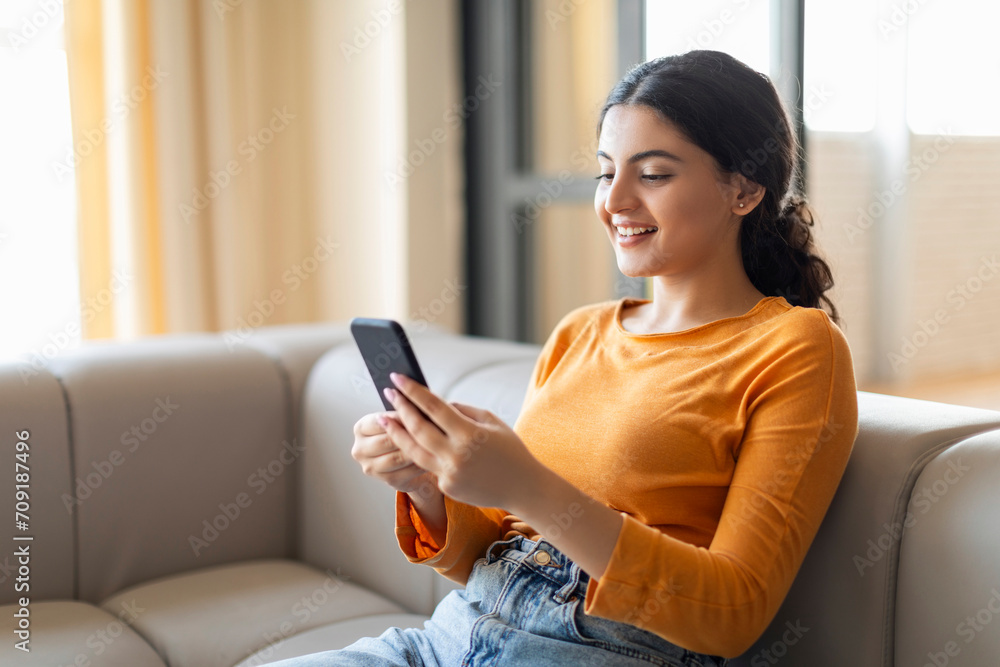 Smiling young indian woman browsing her smartphone while relaxing on couch Smiling young indian woman browsing her smartphone while relaxing on couch