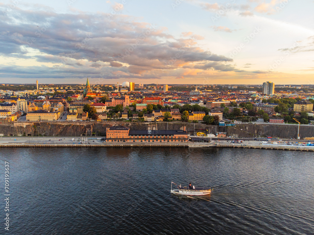 panoramic view over southern Stockholm at sunset, with famous landmarks visible such as the Ericsson Globe. Commuter ferry boat passing by.
