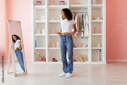 Happy woman trying on oversized jeans at home, mirror reflection