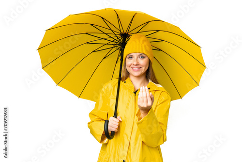 Young blonde woman with rainproof coat and umbrella over isolated chroma key background inviting to come with hand. Happy that you came