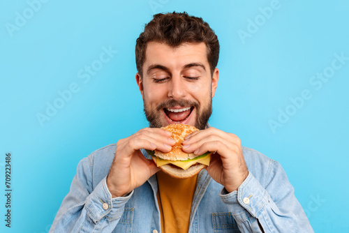Happy caucasian man eats burger and smiling over blue background