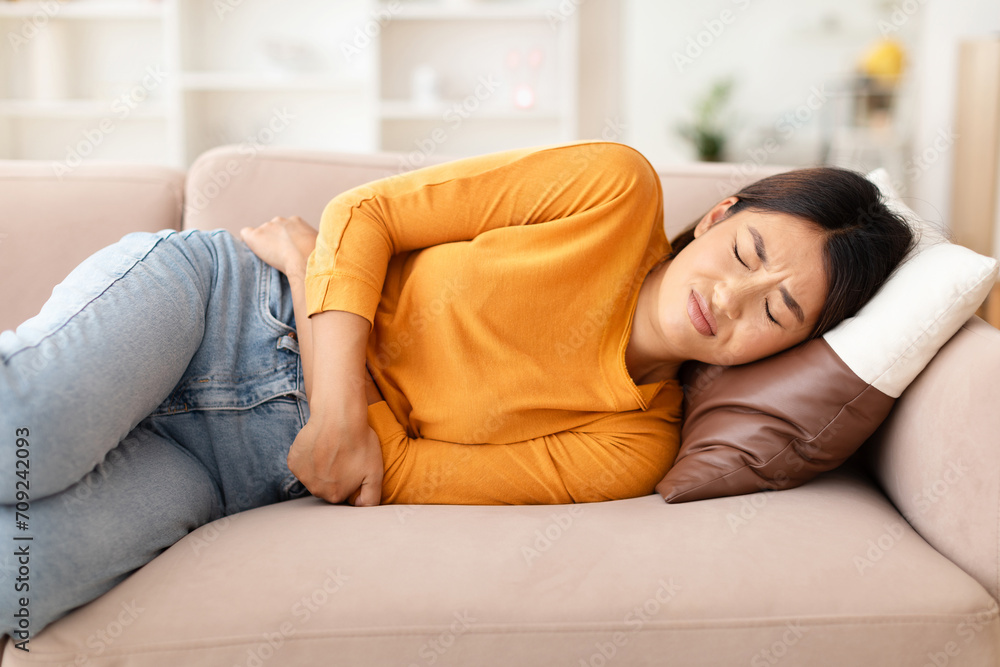 Sick young asian woman laying on couch, touching her belly Sick young asian woman laying on couch, touching her belly