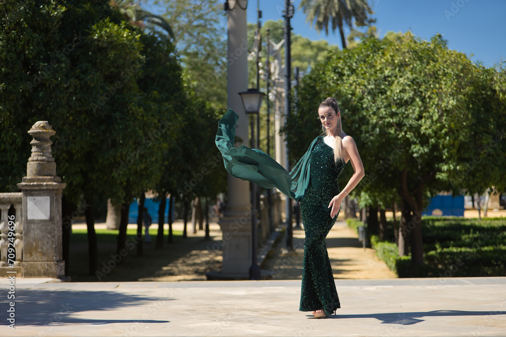 Young, pretty, blonde woman in a green party outfit with sequins, posing as a model while the air blows her cape. Concept of beauty, fashion, trend, model. Young, pretty, blonde woman in a green party outfit with sequins, posing as a model while the air blows her cape. Concept of beauty, fashion, trend, model.