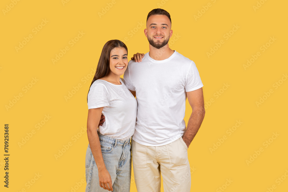 A smiling young couple standing close together in casual clothing against a yellow background A smiling young couple standing close together in casual clothing against a yellow background
