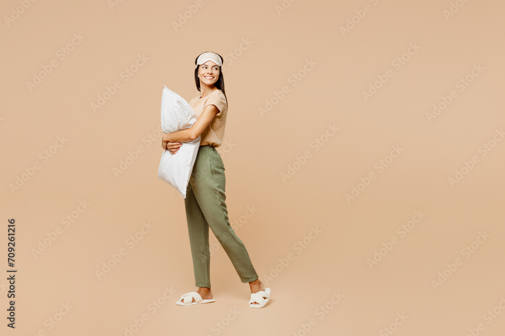 Fototapeta premium Full body side view young calm Latin woman she wears pyjamas jam sleep eye mask rest relax at home hold pillow look aside on area isolated on plain pastel beige background Good mood night nap concept