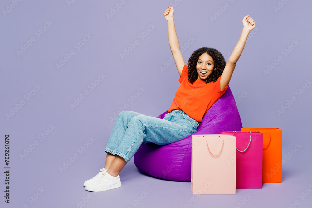 Winner little kid teen girl of African American ethnicity wear orange t-shirt sit in bag chair hold shopping paper package bags isolated on plain purple background. Black Friday sale buy day concept. Winner little kid teen girl of African American ethnicity wear orange t-shirt sit in bag chair hold shopping paper package bags isolated on plain purple background. Black Friday sale buy day concept.