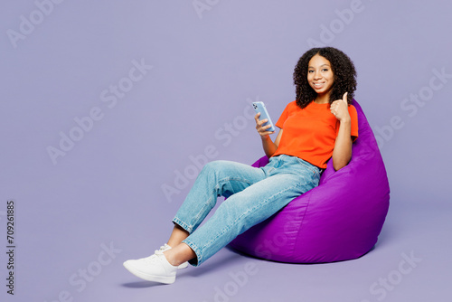 Full body kid teen girl of African American ethnicity wears orange t-shirt sit in bag chair hold use mobile cell phone show thumb up isolated on plain purple background. Childhood lifestyle concept.