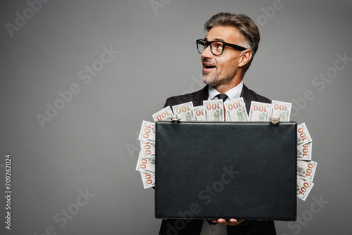 Adult rich employee business man corporate lawyer wearing classic formal black suit shirt tie work in office hold cash money in case for dollar banknotes look aside isolated on plain grey background. photo