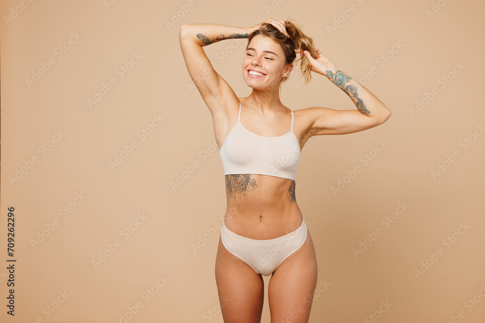 Obraz premium Young calm nice lady woman with slim body perfect skin wear nude top bra lingerie stand looking aside holding hair in hands isolated on plain pastel light beige background. Lifestyle diet fit concept.