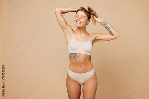 Young calm nice lady woman with slim body perfect skin wear nude top bra lingerie stand looking aside holding hair in hands isolated on plain pastel light beige background. Lifestyle diet fit concept.