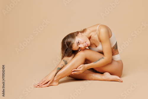 Full body side profile view young nice lady woman with slim body perfect skin wears nude top bra lingerie sit posing hug herself isolated on plain pastel beige background. Lifestyle diet fit concept.