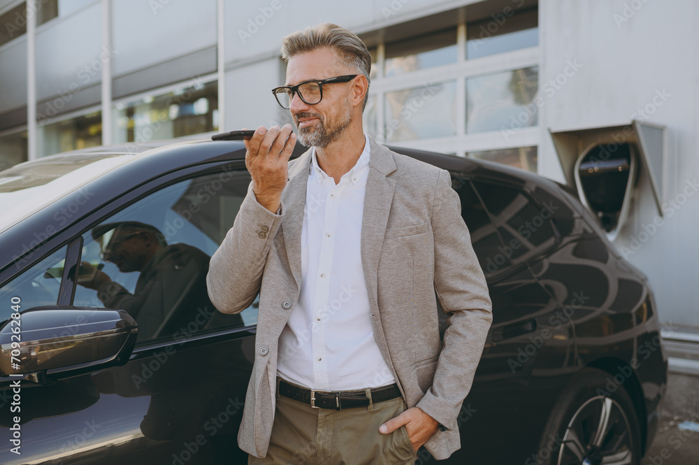Side view adult man customer male buyer client wearing white shirt classic grey suit stand near black electric car while charging record voice message on mobile cell phone outside. Business concept. Side view adult man customer male buyer client wearing white shirt classic grey suit stand near black electric car while charging record voice message on mobile cell phone outside. Business concept.