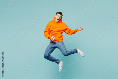 Full body happy singer young man he wears orange hoody casual clothes jump high play do air guitar gesture isolated on plain pastel light blue cyan color background studio portrait. Lifestyle concept.