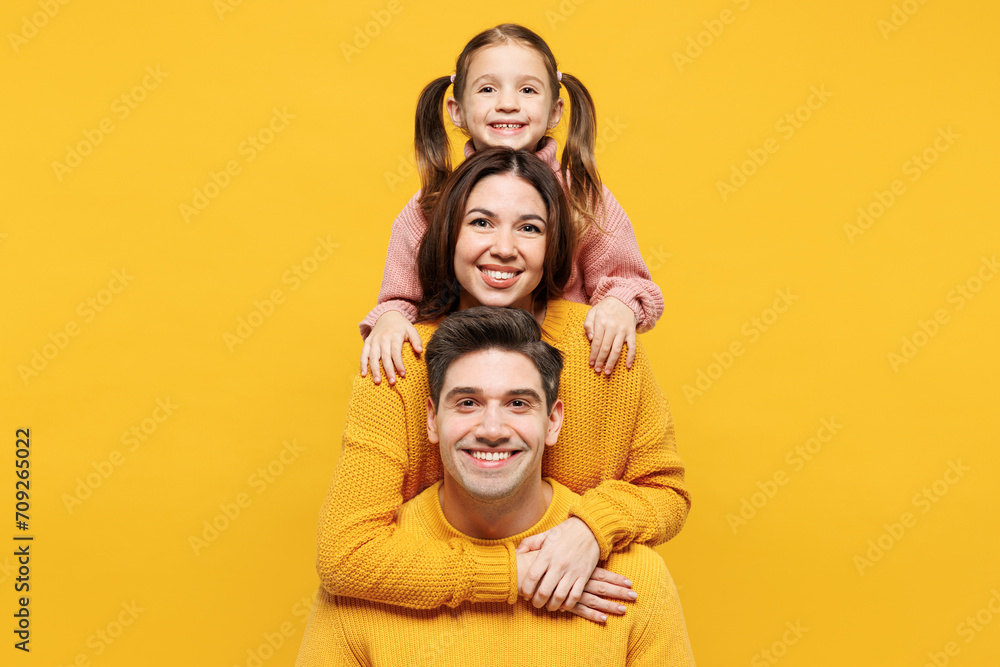Young happy smiling cheerful parents mom dad with child kid girl 7-8 years old wear pink knitted sweater casual clothes stand behind each other isolated on plain yellow background. Family day concept. Young happy smiling cheerful parents mom dad with child kid girl 7-8 years old wear pink knitted sweater casual clothes stand behind each other isolated on plain yellow background. Family day concept.