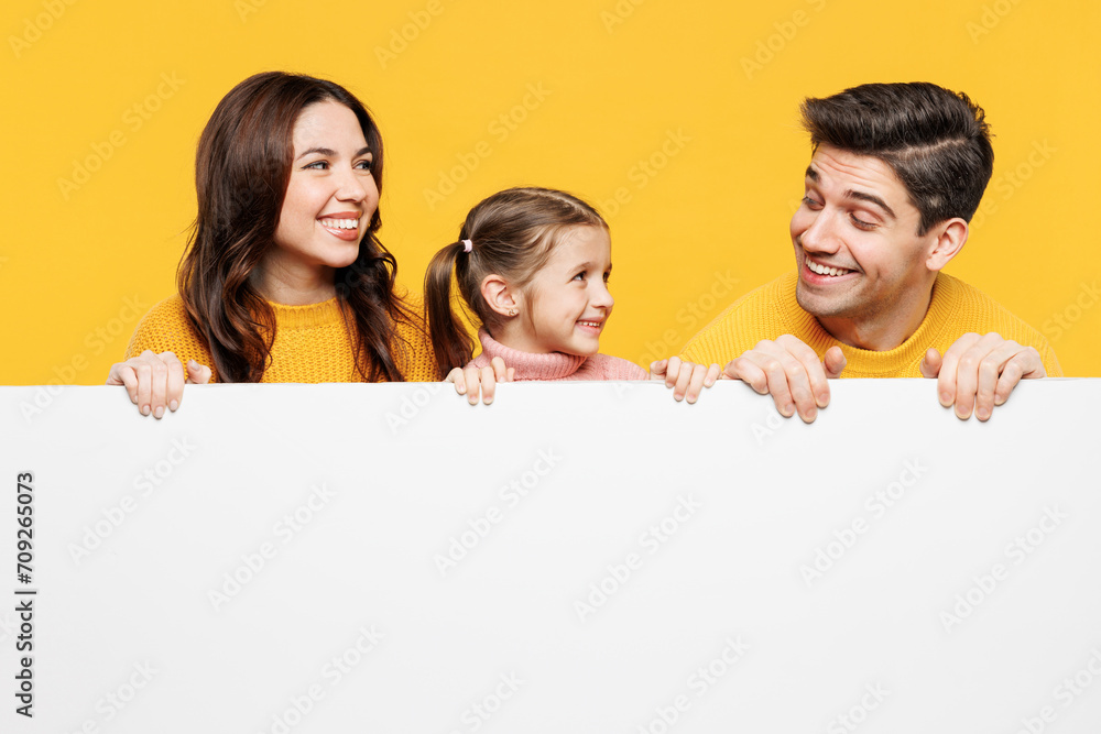 Young parents mom dad with child kid girl 7-8 years old wear pink casual clothes hold big blank poster billboard for content look to each other isolated on plain yellow background. Family day concept. Young parents mom dad with child kid girl 7-8 years old wear pink casual clothes hold big blank poster billboard for content look to each other isolated on plain yellow background. Family day concept.
