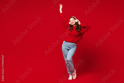 Full body young woman she wearing knitted sweater white hat casual clothes listen to music in headphones dance raise up hand isolated on plain red color background studio portrait. Lifestyle concept.