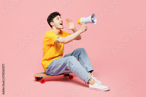 Full body young man wear yellow t-shirt casual clothes sit on skateboard scream in megaphone announces discounts sale Hurry up isolated on plain pastel light pink background studio. Lifestyle concept
