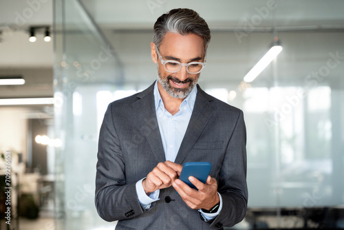 Close-up of smiling mature Latin or Indian businessman holding smartphone in office. Middle aged manager using cell phone mobile app. Digital technology application and solutions for business concept.