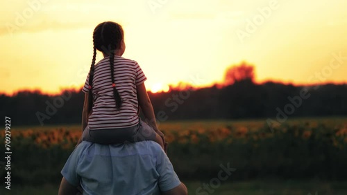 Father carries daughter on shoulders pointing attention to sun on field. Father with daughter on shoulders pointing to far away. Father and daughter on shoulders stretched hands pointing to horizon photo