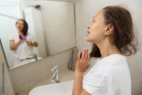 Reflexed young adult brunette woman in mirror combing hair in morning, in the home bathroom