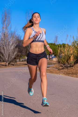 Beautiful tanned young runner girl, dressed in running shorts and white top, running suspended in the air looking at the horizon, smiling happy and free on an asphalt path at sunrise in mountain park. © sirbouman