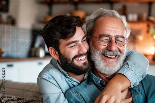An adult son with a beard joyfully hugs his elderly father at home, both smiling and enjoying a relaxed, loving moment together on Father's Day. © sderbane