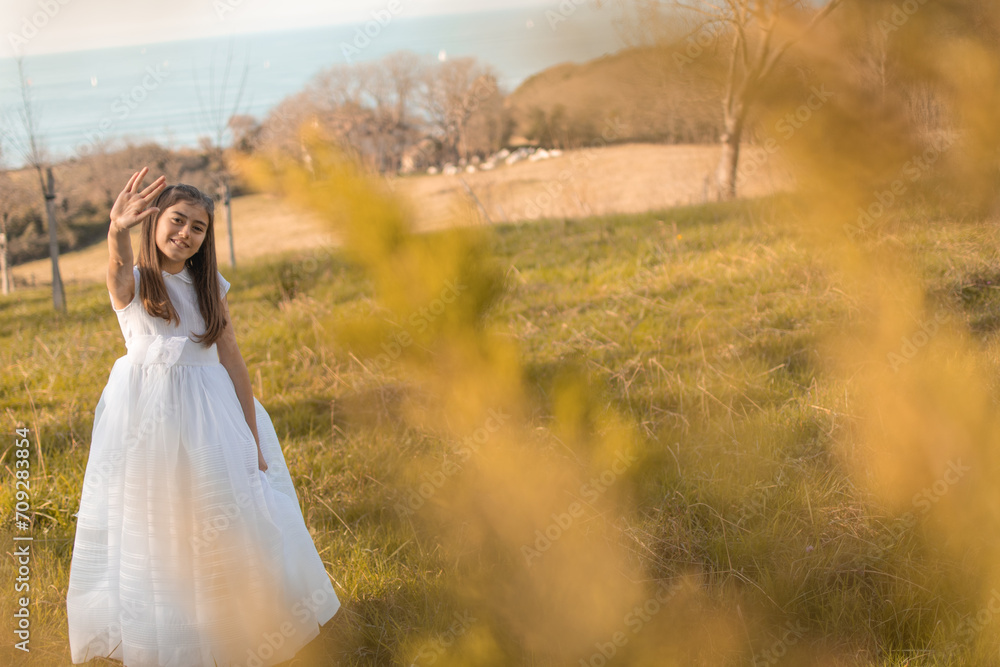 Smiling and adorable girl in a first communion dress in a beautiful field during a warm sunset with the sea in the background Smiling and adorable girl in a first communion dress in a beautiful field during a warm sunset with the sea in the background