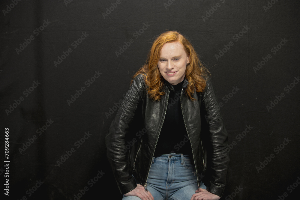 Young woman with red hair in black jacket and jeans poses in studio before a black background Young woman with red hair in black jacket and jeans poses in studio before a black background