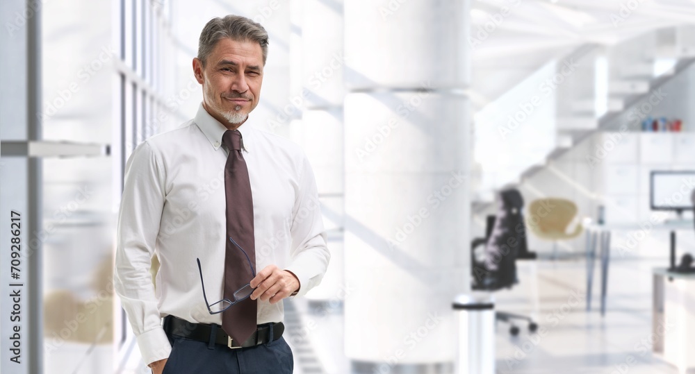 Fototapeta premium Business portrait - happy confident businessman in corporate office, smiling. Mid adult, mature age man standing, looking at camera. Copy space.