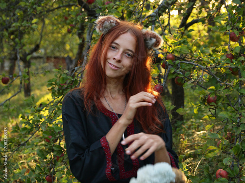 Selfie portraits of a red-haired woman with a fluffy tail and ears in the summer outdoors in an apple orchard. Cosplay for Holo, the wise she-wolf from the anime Spice and the wolf in ethnic clothes.