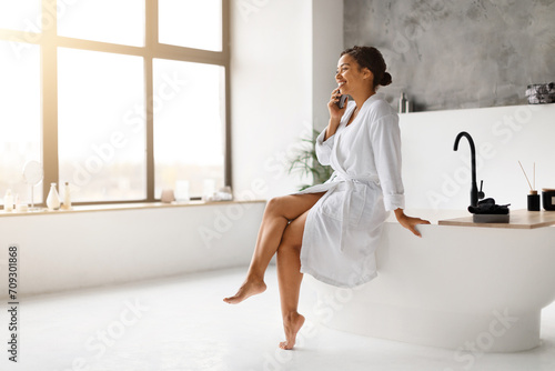 Black woman in bathrobe laughing joyfully while talking on cellphone in bathroom
