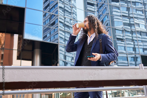 From below mature Latin businessman with long hair enjoying coffee while using a smartphone, in front of modern glass buildings photo