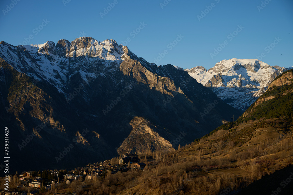 The golden hour illuminates the Pyrenees, casting shadows over the mountainous landscape, with a quaint village nestled at the foothills, in Cerler, Huesca The golden hour illuminates the Pyrenees, casting shadows over the mountainous landscape, with a quaint village nestled at the foothills, in Cerler, Huesca