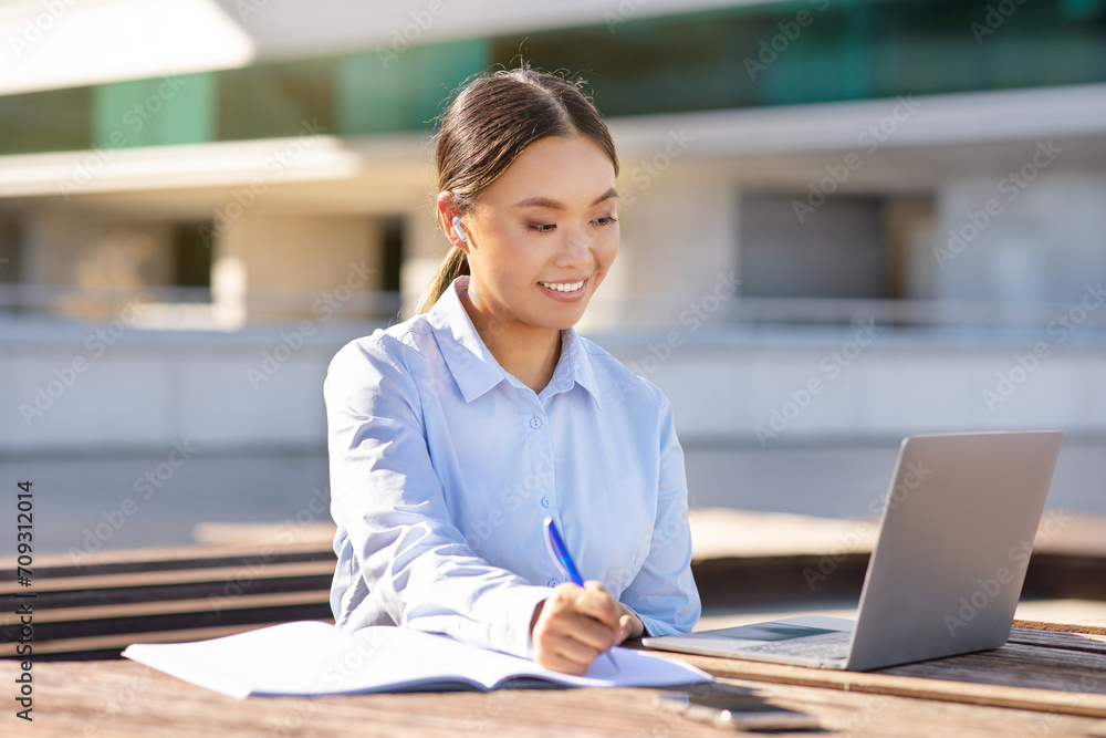 Korean business lady near laptop browsing and writing, sitting outdoors Korean business lady near laptop browsing and writing, sitting outdoors