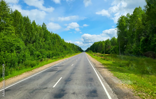A road in a green forest on a summer day. A beautiful straight road, trees with green foliage and a blue sky. A landscape with an empty paved road through the forest in summer © Sofya