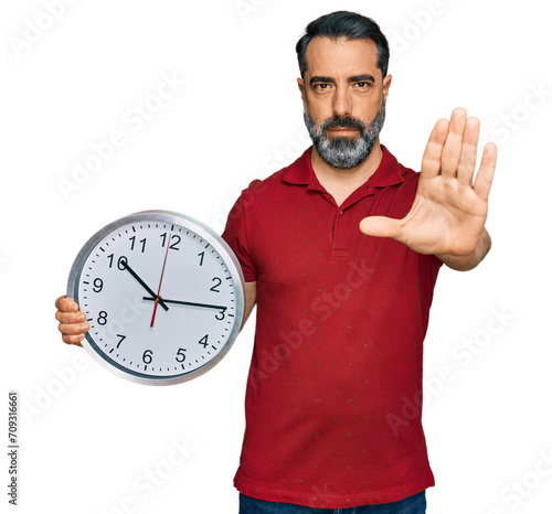 Middle aged man with beard holding big clock with open hand doing stop sign with serious and confident expression, defense gesture