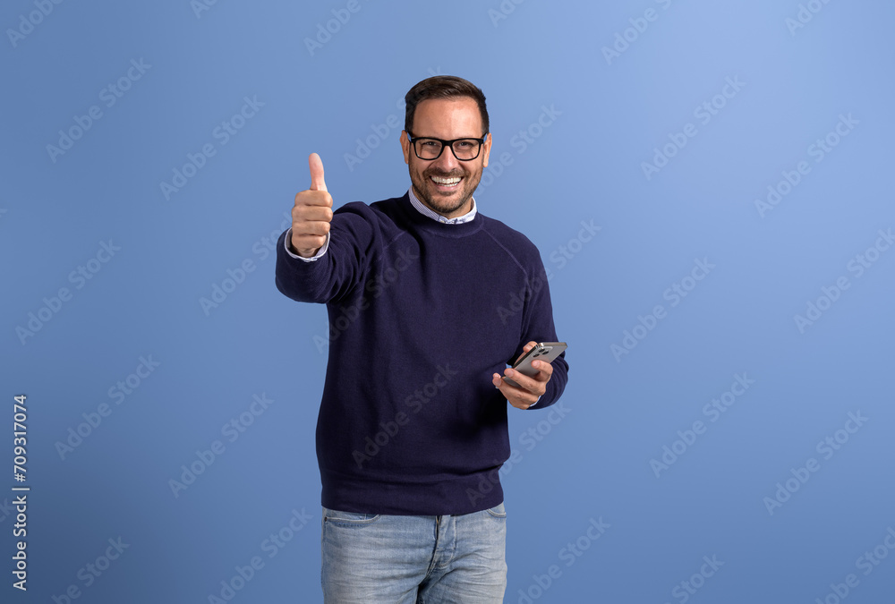 Portrait of confident happy businessman with mobile phone gesturing thumbs up on blue background Portrait of confident happy businessman with mobile phone gesturing thumbs up on blue background