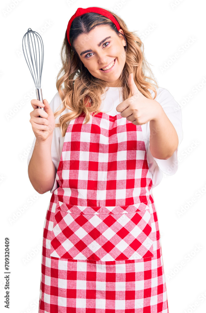 Young caucasian woman wearing apron holding whisk smiling happy and positive, thumb up doing excellent and approval sign Young caucasian woman wearing apron holding whisk smiling happy and positive, thumb up doing excellent and approval sign