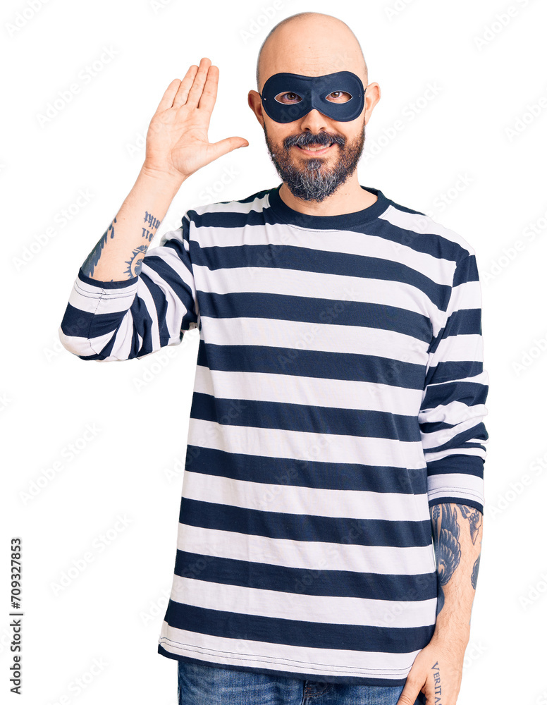 Young handsome man wearing burglar mask waiving saying hello happy and smiling, friendly welcome gesture Young handsome man wearing burglar mask waiving saying hello happy and smiling, friendly welcome gesture