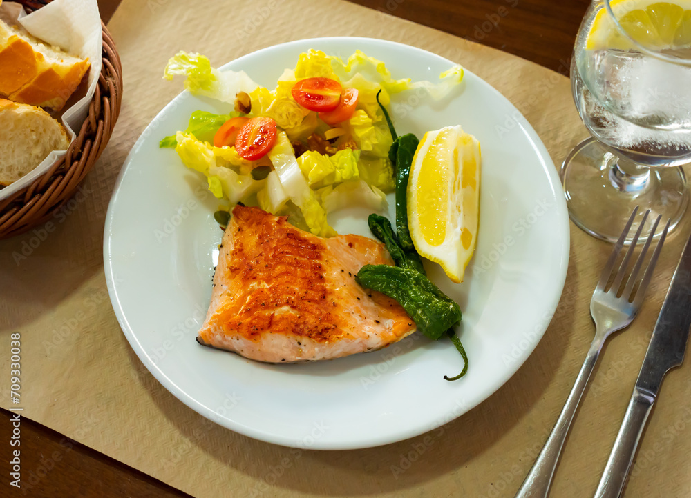 Grilled salmon steak with green pepper, cherry tomatoes, lettuce salad served on table with piece of lemon.