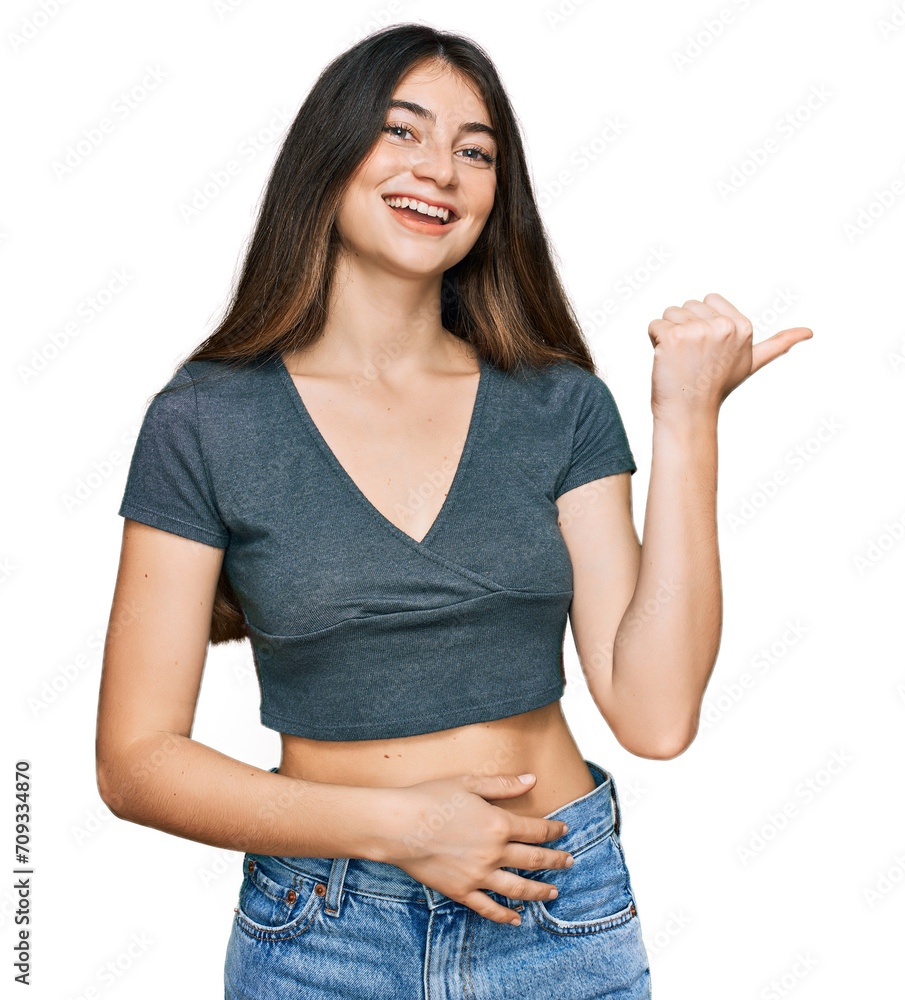 Young beautiful teen girl wearing casual crop top t shirt smiling with happy face looking and pointing to the side with thumb up. Young beautiful teen girl wearing casual crop top t shirt smiling with happy face looking and pointing to the side with thumb up.