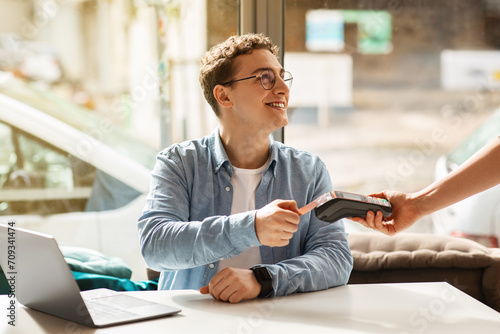 Cheerful young european man using credit card for payment machine, sit on desk with laptop photo