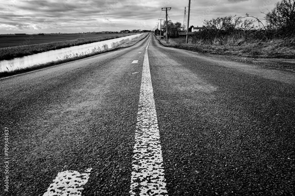 Ground level view of a dangerous fenland road showing the road sinking in places. A deep river is on the left, the road has had serious accidents over the years.
