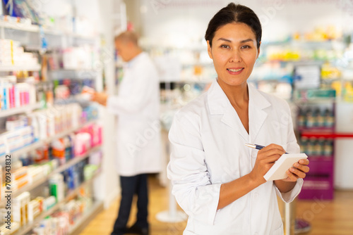 Female Asian pharmacist in white gown standing in salesroom of chemist shop with pen and sheets of paper in hands and looking in camera. photo