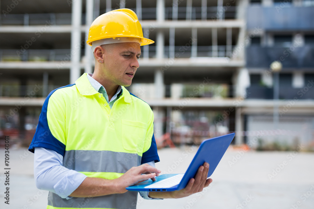 Portrait of positive man architect standing with laptop in construction site outdoors