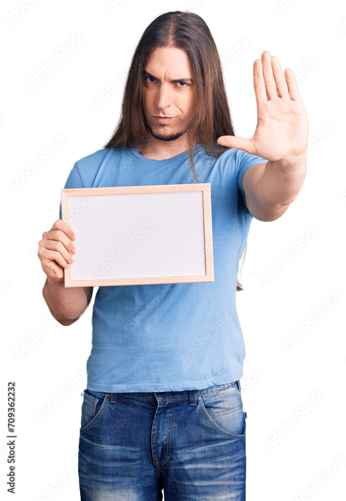 Young adult man with long hair holding empty white chalkboard with open hand doing stop sign with serious and confident expression, defense gesture Young adult man with long hair holding empty white chalkboard with open hand doing stop sign with serious and confident expression, defense gesture