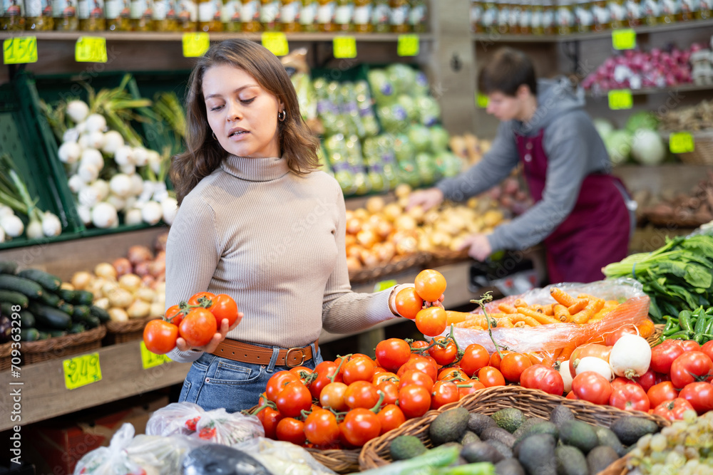 Positive young girl choosing tomatoes standing at counter in large vegetable market