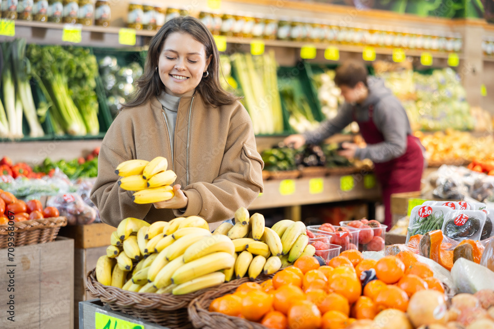 Young woman shopper in casual clothes chooses bananas in vegetable shop